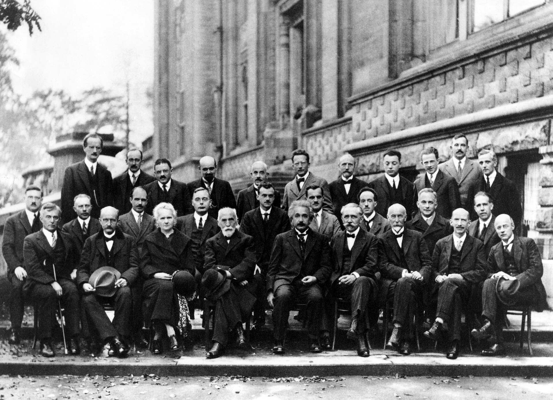 Fotografía en blanco y negro de 29 científicos (28 hombres y una mujer) posando en tres filas frente a un edificio clásico de piedra con ventanas grandes. La primera fila está sentada en sillas, mientras que las dos filas traseras están de pie. Todos visten trajes formales oscuros de la década de 1920. En el centro de la fila sentada se distingue a Marie Curie.